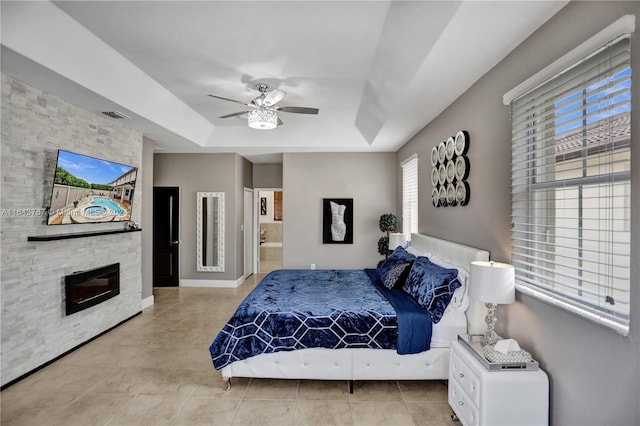 bedroom with ensuite bath, a raised ceiling, ceiling fan, a stone fireplace, and light tile patterned flooring