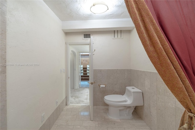 half bath with a textured ceiling, toilet, visible vents, tile walls, and wainscoting