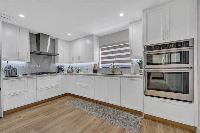 kitchen with stainless steel appliances, wall chimney exhaust hood, sink, light wood-type flooring, and white cabinets