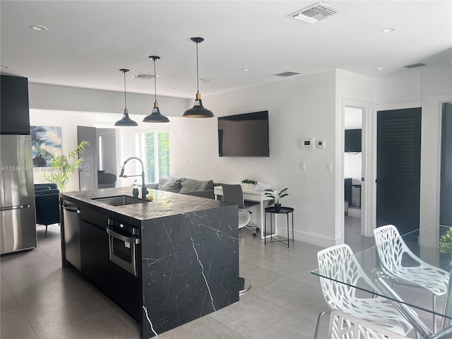 kitchen with hanging light fixtures, stone countertops, stainless steel appliances, a center island with sink, and sink