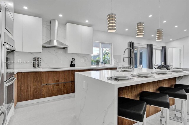 kitchen with white cabinetry, tasteful backsplash, a kitchen island with sink, wall chimney range hood, and light tile patterned floors