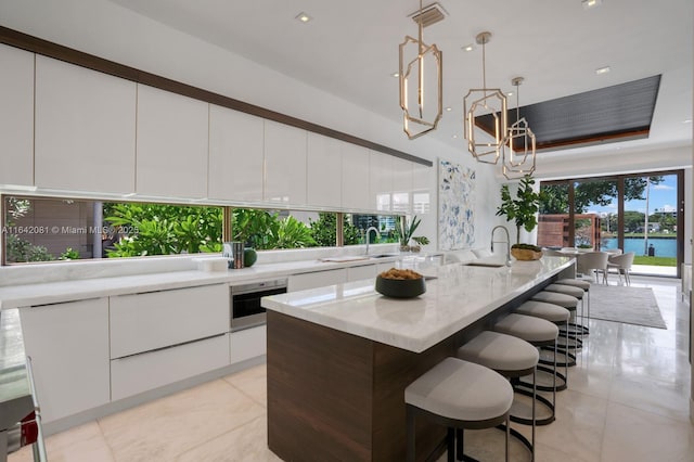 kitchen with light stone countertops, pendant lighting, a water view, white cabinets, and an island with sink