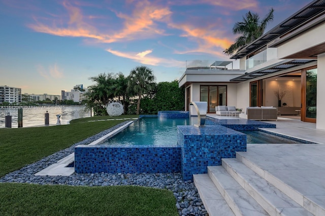 pool at dusk with a yard, an outdoor living space, a hot tub, pool water feature, and a water view