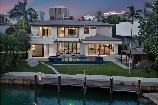 back house at dusk featuring a patio area, a balcony, a swimming pool with hot tub, and a water view