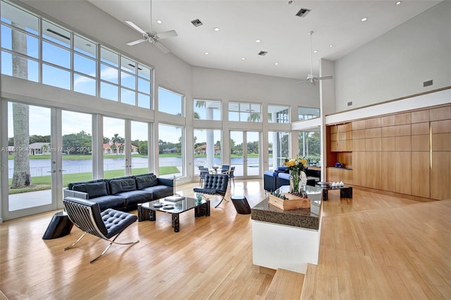 living room featuring ceiling fan, light hardwood / wood-style flooring, a towering ceiling, french doors, and a water view