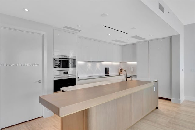 kitchen featuring decorative backsplash, light wood-type flooring, stainless steel double oven, a kitchen island with sink, and white cabinets