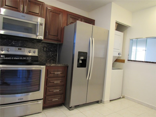 kitchen with stacked washer / drying machine, dark stone countertops, light tile patterned floors, appliances with stainless steel finishes, and backsplash