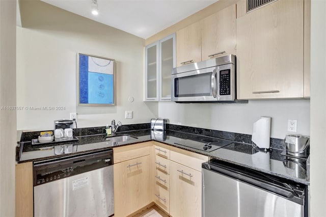 kitchen with light brown cabinetry, appliances with stainless steel finishes, glass insert cabinets, a sink, and dark stone countertops