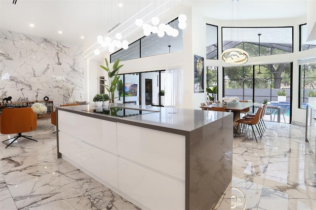 kitchen with an island with sink, white cabinetry, black electric cooktop, pendant lighting, and a towering ceiling