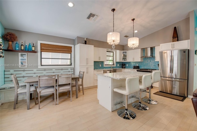 kitchen with wall chimney exhaust hood, hanging light fixtures, white cabinetry, light stone countertops, and stainless steel fridge