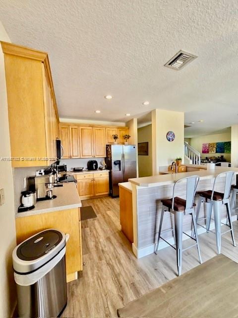 kitchen with appliances with stainless steel finishes, a breakfast bar, a textured ceiling, light brown cabinetry, and light hardwood / wood-style flooring