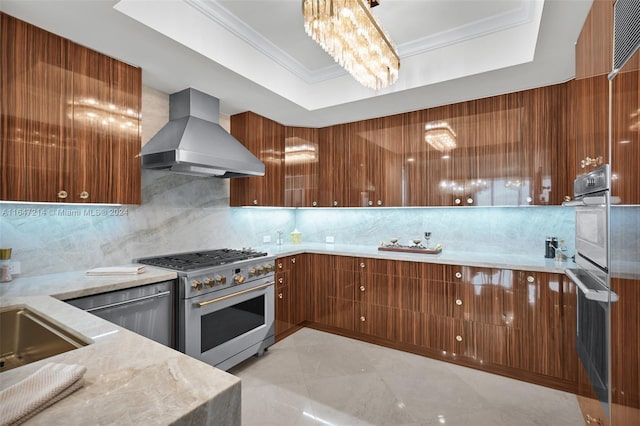 kitchen featuring a tray ceiling, wall chimney range hood, stainless steel appliances, backsplash, and ornamental molding