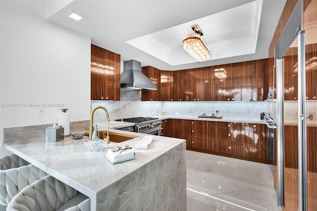 kitchen featuring high end stainless steel range oven, sink, kitchen peninsula, wall chimney range hood, and a raised ceiling