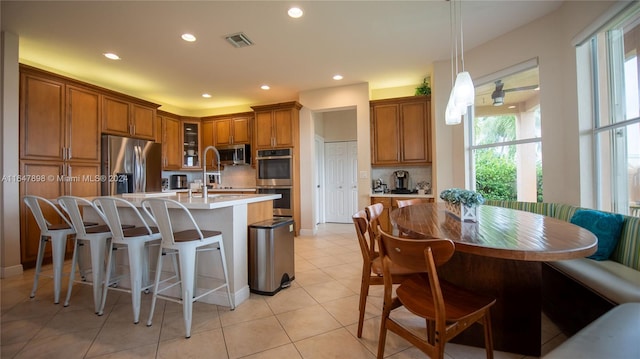 kitchen featuring a kitchen island with sink, backsplash, light tile patterned floors, decorative light fixtures, and appliances with stainless steel finishes