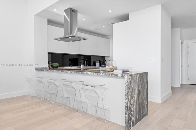 kitchen with light wood-type flooring, kitchen peninsula, light stone countertops, island exhaust hood, and white cabinets