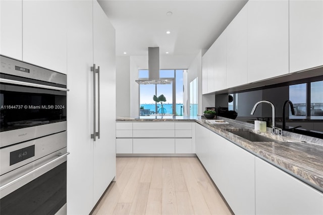 kitchen featuring light wood-style floors, stainless steel double oven, a sink, stone counters, and white cabinetry