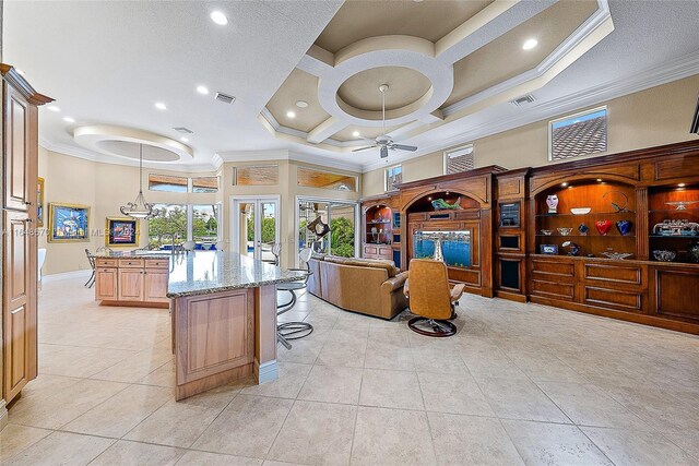 kitchen with coffered ceiling, ornamental molding, light stone counters, ceiling fan, and a breakfast bar area