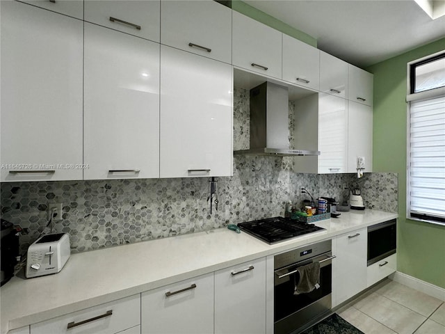 kitchen featuring backsplash, wall chimney exhaust hood, light tile patterned floors, stainless steel appliances, and white cabinets