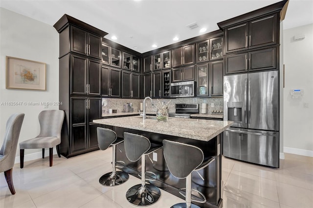 kitchen with light tile patterned floors, light stone counters, stainless steel appliances, an island with sink, and decorative backsplash