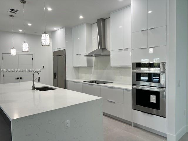 kitchen with pendant lighting, wall chimney range hood, sink, white cabinetry, and a kitchen island with sink