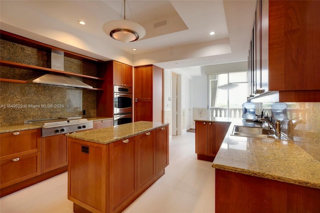 kitchen with a raised ceiling, stainless steel appliances, a kitchen island, sink, and wall chimney exhaust hood