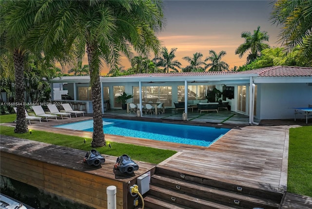 pool at dusk with outdoor lounge area, a deck, a patio, and a lawn