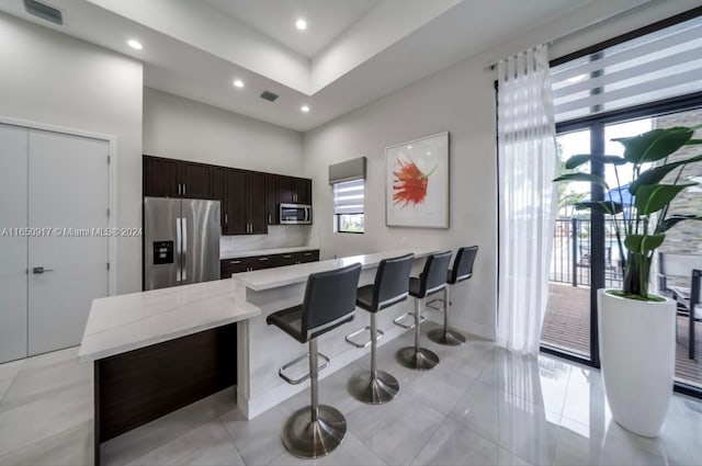 kitchen featuring backsplash, stainless steel appliances, dark brown cabinets, light tile patterned flooring, and a kitchen bar