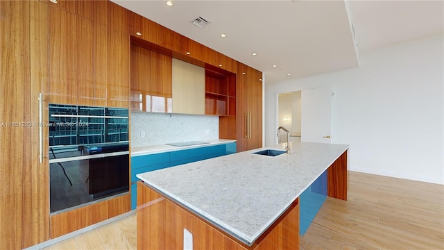 kitchen featuring light stone counters, sink, black electric cooktop, a center island with sink, and light hardwood / wood-style floors