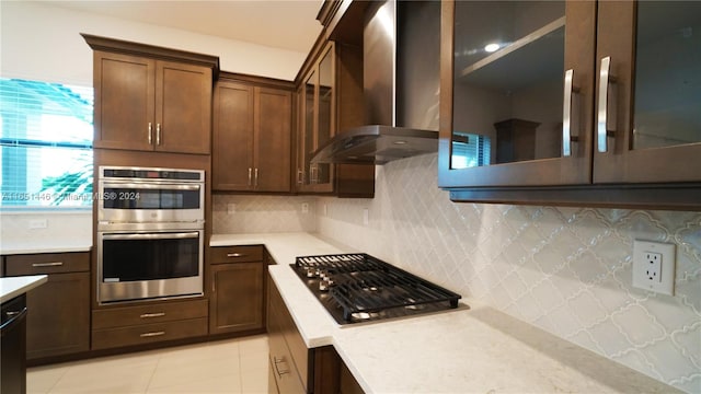 kitchen featuring light tile patterned floors, stainless steel appliances, decorative backsplash, wall chimney exhaust hood, and dark brown cabinetry