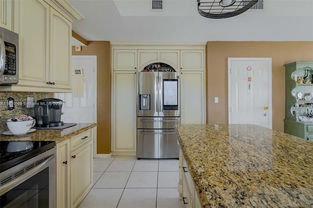 kitchen with cream cabinets, light tile patterned floors, stainless steel appliances, decorative backsplash, and light stone counters