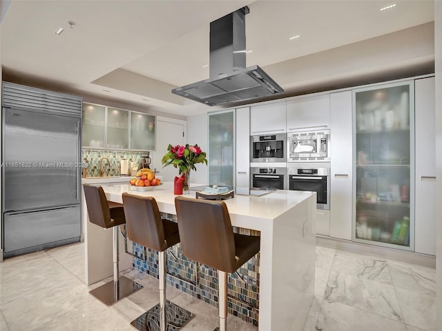 kitchen featuring a center island, built in appliances, a kitchen breakfast bar, island range hood, and tasteful backsplash