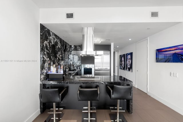 kitchen with a kitchen breakfast bar, dark stone countertops, island range hood, kitchen peninsula, and stainless steel oven