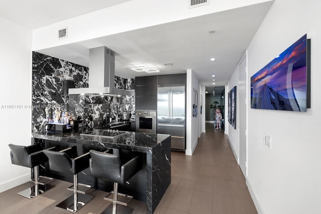 kitchen featuring dark stone counters, tile walls, kitchen peninsula, wall chimney range hood, and stainless steel appliances