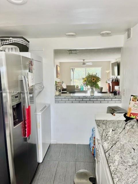 kitchen with light stone counters, white cabinetry, stacked washing maching and dryer, ceiling fan, and stainless steel fridge