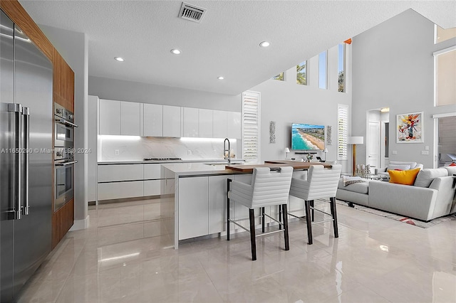 kitchen featuring appliances with stainless steel finishes, sink, white cabinetry, a high ceiling, and a center island with sink