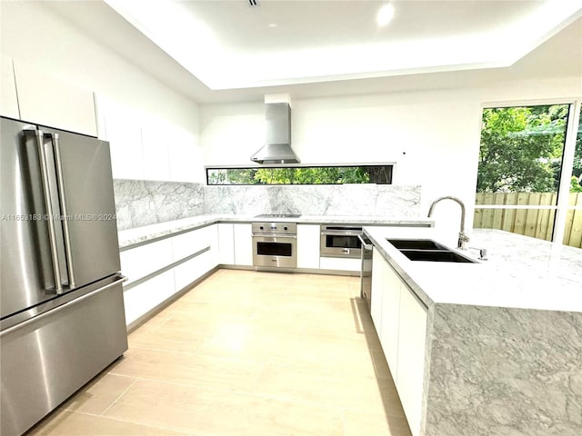 kitchen featuring light stone countertops, stainless steel appliances, white cabinets, sink, and wall chimney range hood