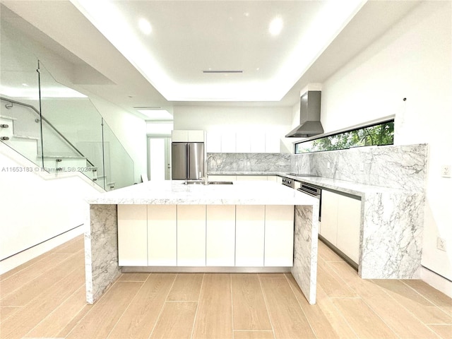 kitchen featuring light wood-type flooring, wall chimney range hood, stainless steel refrigerator, and white cabinets