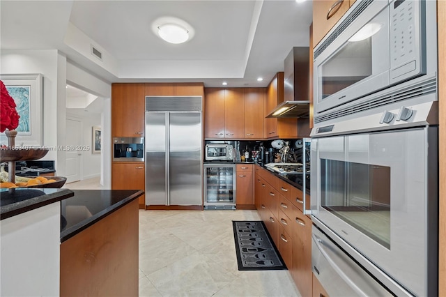 kitchen with wine cooler, wall chimney exhaust hood, built in appliances, and dark stone countertops