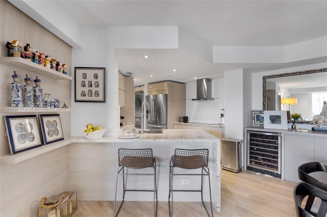 kitchen featuring light wood-type flooring, wall chimney range hood, stainless steel fridge, beverage cooler, and a breakfast bar area