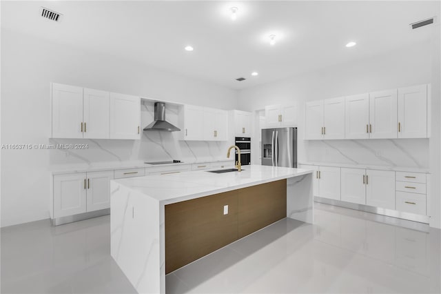 kitchen featuring backsplash, light stone counters, stainless steel fridge, wall chimney exhaust hood, and an island with sink