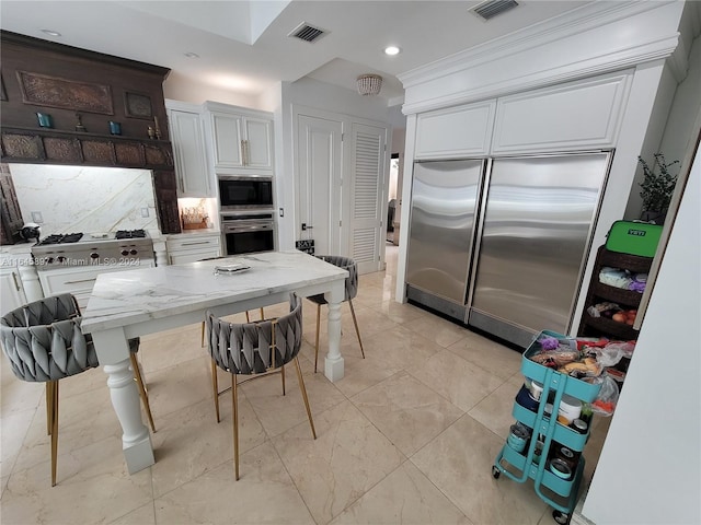 kitchen with light stone counters, ornamental molding, backsplash, white cabinetry, and built in appliances