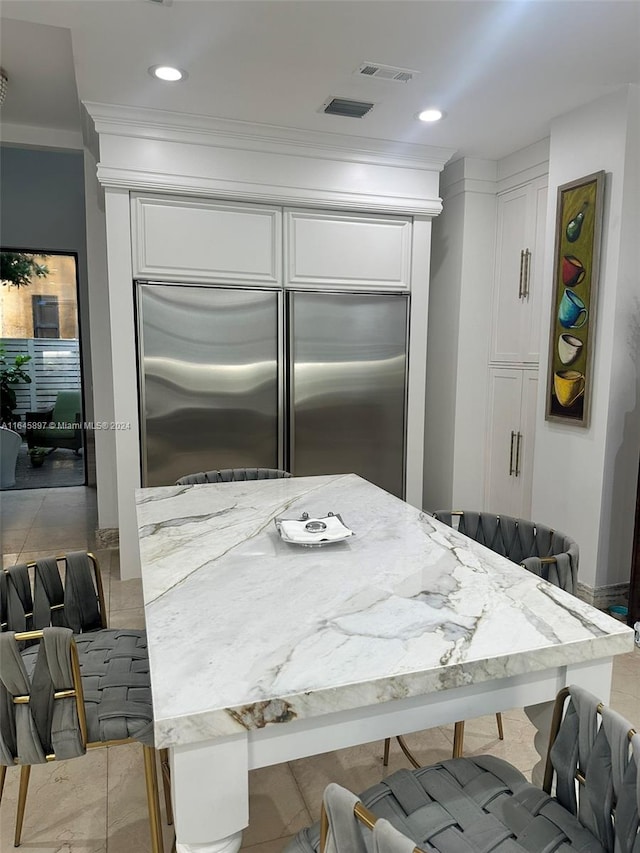 kitchen with ornamental molding, built in refrigerator, light stone counters, and white cabinetry