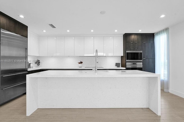 kitchen featuring light wood-type flooring, built in appliances, dark brown cabinets, and an island with sink