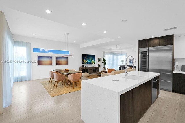 kitchen with a kitchen island with sink, light hardwood / wood-style flooring, sink, stainless steel built in refrigerator, and dark brown cabinetry