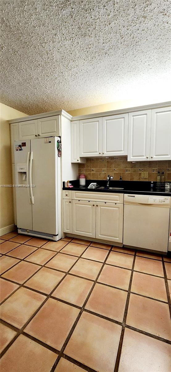 kitchen featuring a textured ceiling, white appliances, light tile patterned floors, backsplash, and white cabinets