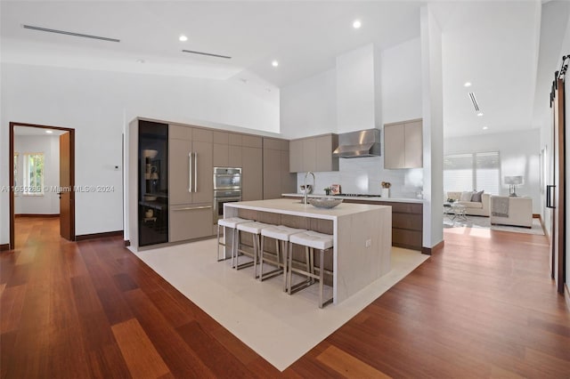 kitchen featuring a kitchen breakfast bar, wall chimney exhaust hood, dark wood-type flooring, a center island with sink, and high vaulted ceiling