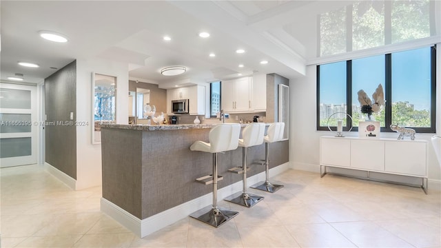 kitchen featuring a kitchen bar, kitchen peninsula, white cabinetry, stone counters, and light tile patterned flooring