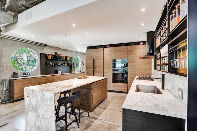 kitchen featuring a kitchen island, light stone countertops, a breakfast bar, wall chimney range hood, and double oven