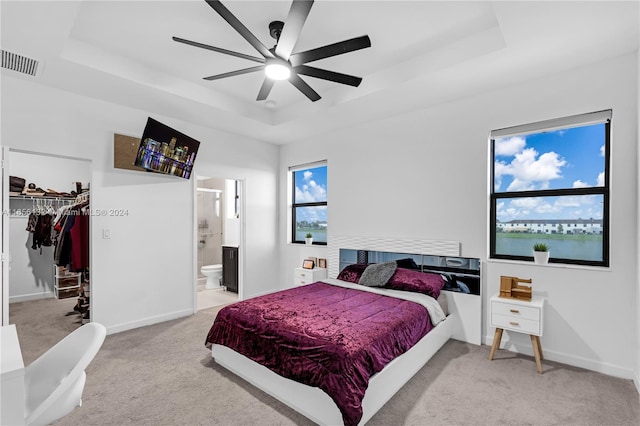 carpeted bedroom featuring a raised ceiling, ceiling fan, a walk in closet, and ensuite bath