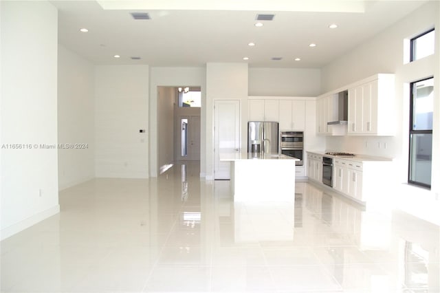 kitchen featuring appliances with stainless steel finishes, wall chimney exhaust hood, a kitchen island with sink, light tile patterned floors, and white cabinetry